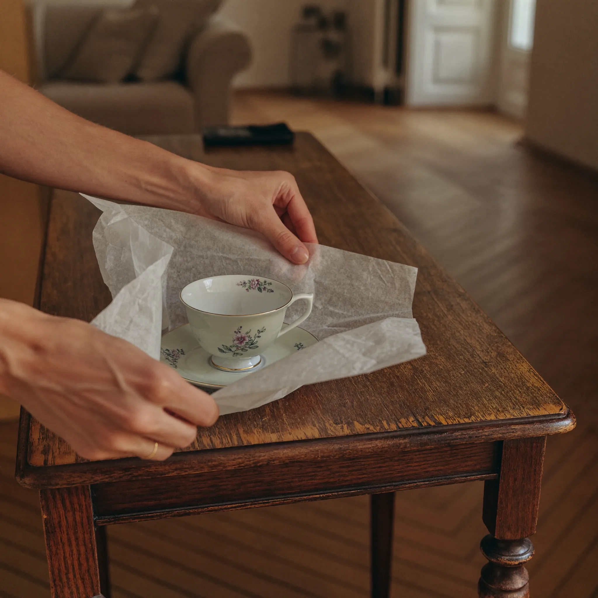 Hände verpacken sorgfältig eine Porzellantasse in Seidenpapier auf einem alten Holztisch in einer Altbauwohnung
